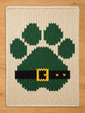 A rectangular chunky knit blanket in cream featuring a large green paw print design. The paw print includes a black horizontal band across the bottom section with a yellow rectangular buckle detail in the center of the band.