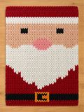 A rectangular chunky knit blanket featuring a large, stylized Santa Claus pattern. The knitted design shows a red hat with a white brim, a peach face with black eyes and a round pink nose, a large white V-shaped beard, and a red suit with a black belt and yellow buckle, laid flat on a light wooden surface