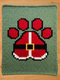 A knitted green chunky blanket with a Santa Claus paw print design. The paw print is red with black and white accents, and a gold buckle. The blanket is made of a soft, textured material.