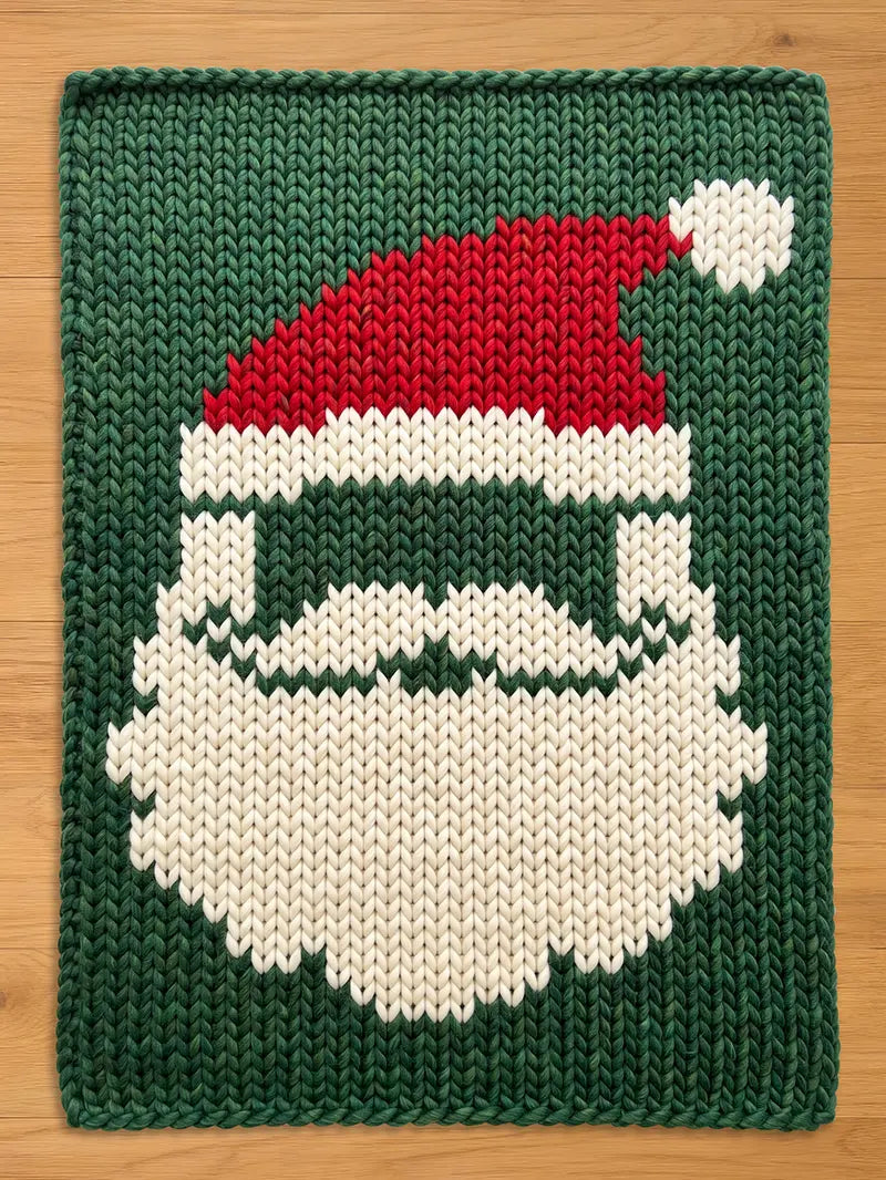 A knitted Christmas-themed chunky blanket featuring a Santa Claus face. The design includes a red hat with a white pom-pom, a white beard, and a green background. The blanket is rectangular.