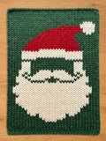 A knitted Christmas-themed chunky blanket featuring a Santa Claus face. The design includes a red hat with a white pom-pom, a white beard, and a green background. The blanket is rectangular.