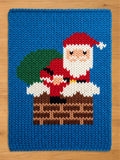 A knitted blue Christmas-themed blanket featuring Santa Claus in a chimney. Santa is wearing a red suit and hat, with a white beard and a green sack of gifts. The chimney is brown, and the blanket has a blue border.