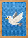 A knitted chunky blanket in a vibrant blue color, featuring a white seagull design with gray and yellow accents. The seagull is depicted in flight, adding a dynamic element to the textile art. The piece is likely a decorative item or a baby blanket.