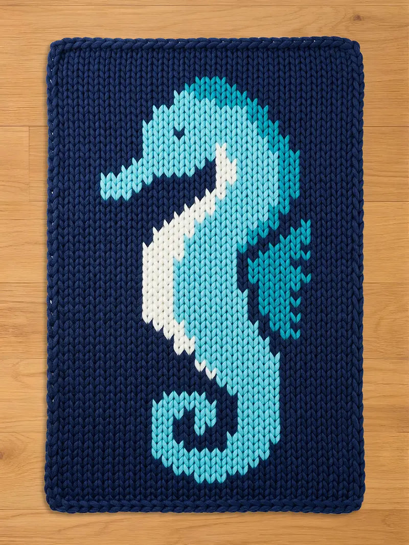 A navy blue knitted chunky blanket featuring a seahorse design. The seahorse is rendered in shades of light blue, white, and teal. The blanket is made of a thick, textured yarn, suitable for a nursery or children's room.