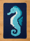 A navy blue knitted chunky blanket featuring a seahorse design. The seahorse is rendered in shades of light blue, white, and teal. The blanket is made of a thick, textured yarn, suitable for a nursery or children's room.