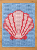 A chunky knit blanket laid flat on a wooden surface. It features a light blue background with a large, stylized pink and coral seashell pattern knitted into the center