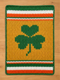 A knitted rectangular chunky blanket featuring a large green shamrock on a yellow background. The blanket has green borders and stripes of white, orange, and green at the top and bottom. The design is reminiscent of Irish heritage.