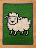 Chunky knit blanket featuring a stylized white sheep with black outlines on a vibrant green background, finished with a dark blue border.