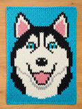 A knitted chunky blanket featuring a smiling husky dog face. The dog has black and white fur, blue eyes, and a pink tongue. The background is a bright blue, and the piece is likely a blanket or pillow.