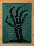A rectangular knitted textile featuring a chunky knit texture in a deep teal green color. The center of the piece displays a bold, black graphic design of a stylized, leafless tree with branches reaching upward from a solid base.