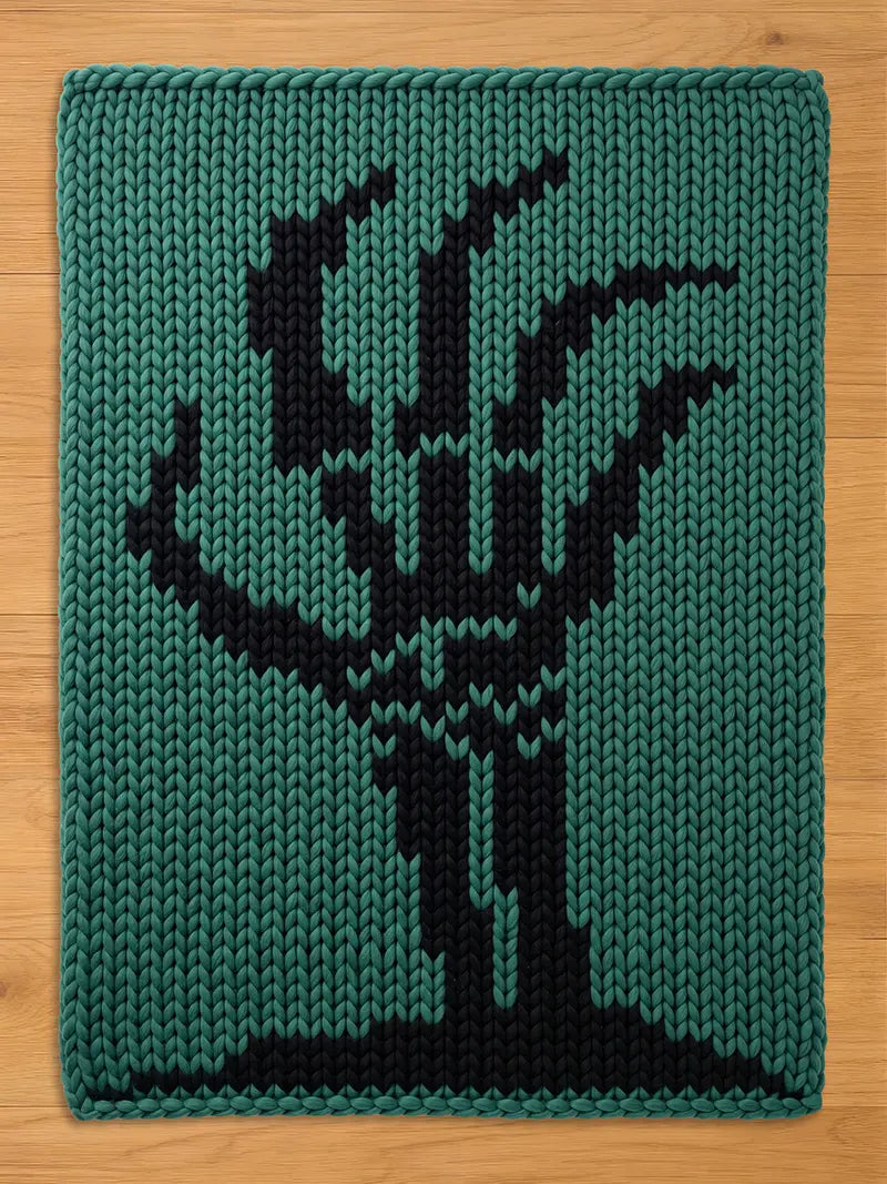 A rectangular knitted textile featuring a chunky knit texture in a deep teal green color. The center of the piece displays a bold, black graphic design of a stylized, leafless tree with branches reaching upward from a solid base.
