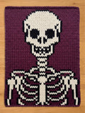 A chunky knit blanket featuring a large white and black skeleton design, including a skull and ribcage, set against a deep purple background
