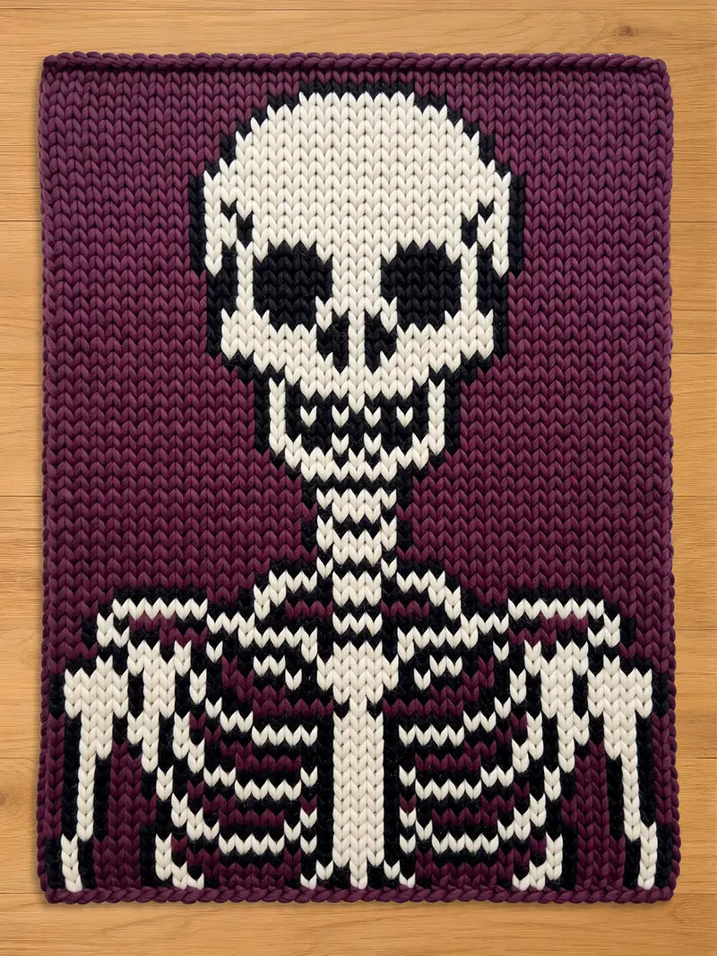 A chunky knit blanket featuring a large white and black skeleton design, including a skull and ribcage, set against a deep purple background