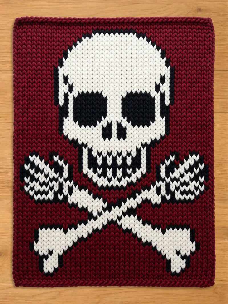 A cozy, deep burgundy chunky knit blanket featuring a graphic white skull and crossbones pattern with skeleton hands. The skull details are accented with black knit stitches