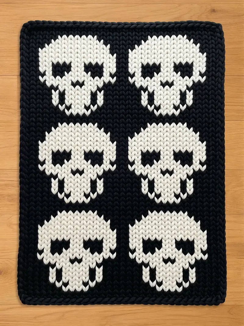 A rectangular chunky knit blanket laid flat on a light wood surface. The blanket has a solid black background with a repeating pattern of six large, stylized white skulls arranged in a grid of two columns and three rows