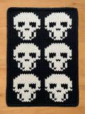 A rectangular chunky knit blanket laid flat on a light wood surface. The blanket has a solid black background with a repeating pattern of six large, stylized white skulls arranged in a grid of two columns and three rows