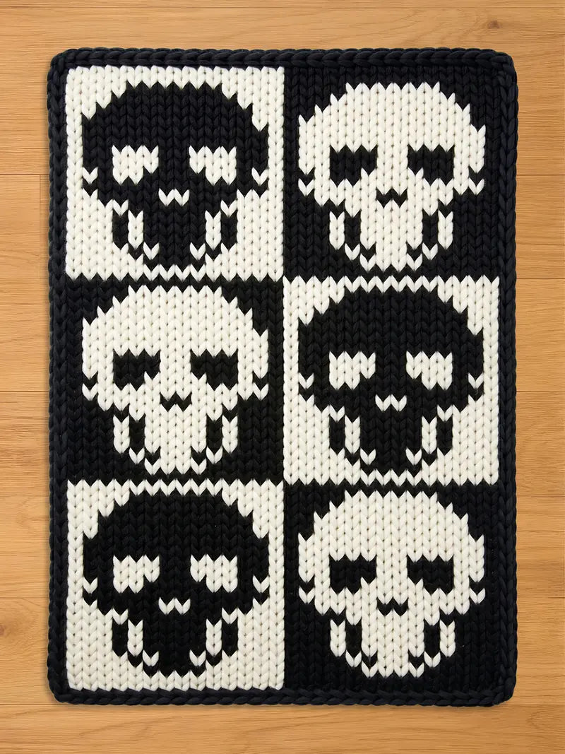 A rectangular chunky knit blanket featuring a bold, high-contrast gothic design. Six stylized skulls are arranged in a two-by-three checkerboard grid, alternating between black skulls on a white background and white skulls on a black background, framed by a solid black knitted edge