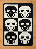 A rectangular chunky knit blanket featuring a bold, high-contrast gothic design. Six stylized skulls are arranged in a two-by-three checkerboard grid, alternating between black skulls on a white background and white skulls on a black background, framed by a solid black knitted edge