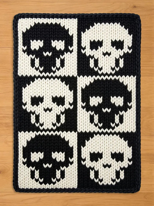 A rectangular chunky knit blanket featuring a bold, high-contrast gothic design. Six stylized skulls are arranged in a two-by-three checkerboard grid, alternating between black skulls on a white background and white skulls on a black background, framed by a solid black knitted edge