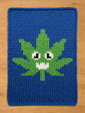 A deep blue chunky knit blanket laid flat on a light wooden surface, featuring a bright green, stylized leaf in the center. The leaf has a playful cartoon face with large black and white eyes and a jagged white smile