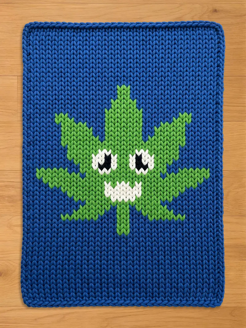 A deep blue chunky knit blanket laid flat on a light wooden surface, featuring a bright green, stylized leaf in the center. The leaf has a playful cartoon face with large black and white eyes and a jagged white smile