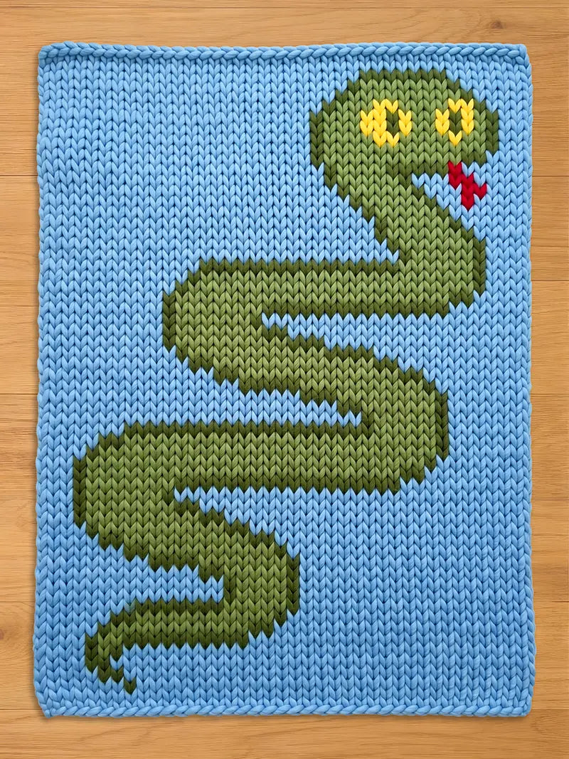  A knitted chunky blanket featuring a green snake design on a light blue background. The snake has yellow eyes and a red tongue. The knitted texture is visible throughout the design, creating a handmade aesthetic.