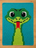A rectangular knitted blanket featuring a large, centered snake head design. The snake is rendered in shades of lime and forest green with black and white eyes and a bright red forked tongue, all set against a vibrant teal background.