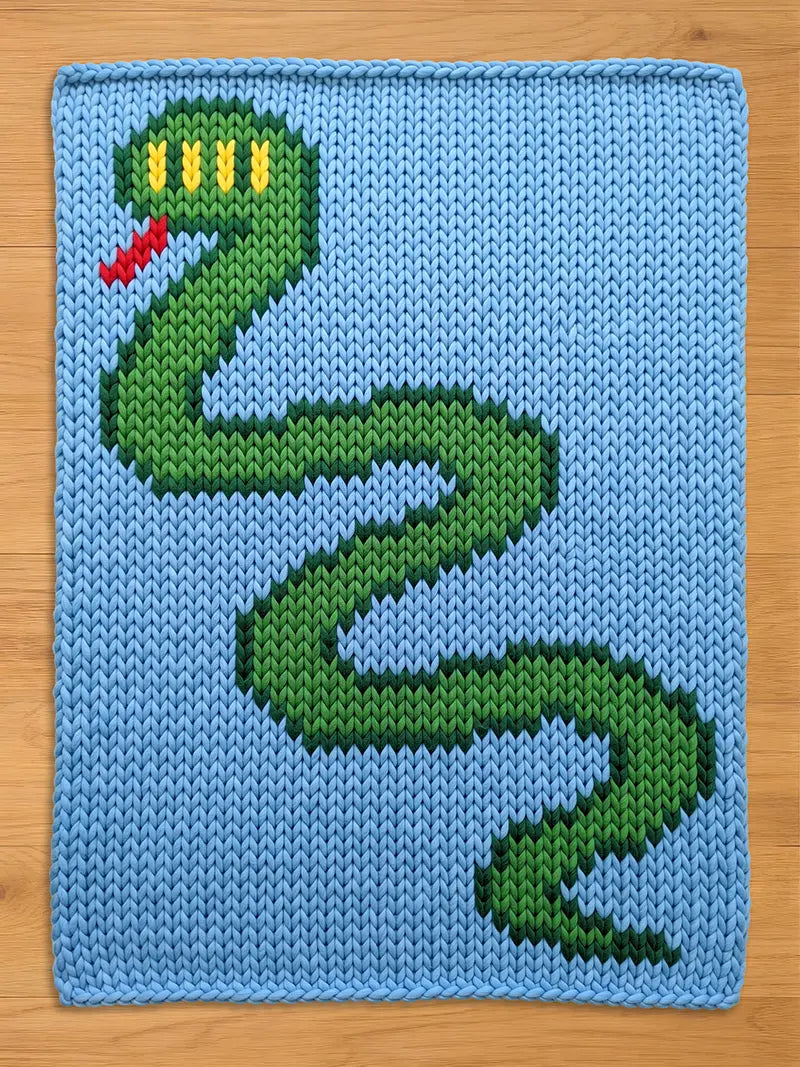 A knitted chunky blanket featuring a green snake design on a light blue background. The snake has a green body, yellow eyes, and a red tongue. The piece is made of yarn.