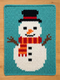 A rectangular knit blanket featuring a white snowman on a teal background. The snowman wears a black top hat with a red band, an orange carrot nose, and a red and orange striped scarf. Three black buttons are on the snowman's torso.