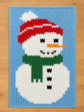 A knitted snowman design on a blue background. The snowman has a red and white hat, a green scarf, and a carrot nose. Black buttons and eyes complete the design. The background is a light blue color.