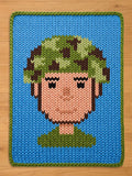 A knitted chunky blanket featuring a pixelated face with a green leafy hair design. The face is peach-colored with black eyes and a green collar. The blanket has a blue background with a green border.
