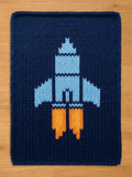 Dark blue chunky knit blanket featuring a large light blue spaceship design with bright orange exhaust flames.