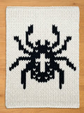 A square, off-white knitted blanket featuring a large, black pixelated insect design. The insect has a segmented body, multiple legs, and antennae. The blanket is made of thick yarn.