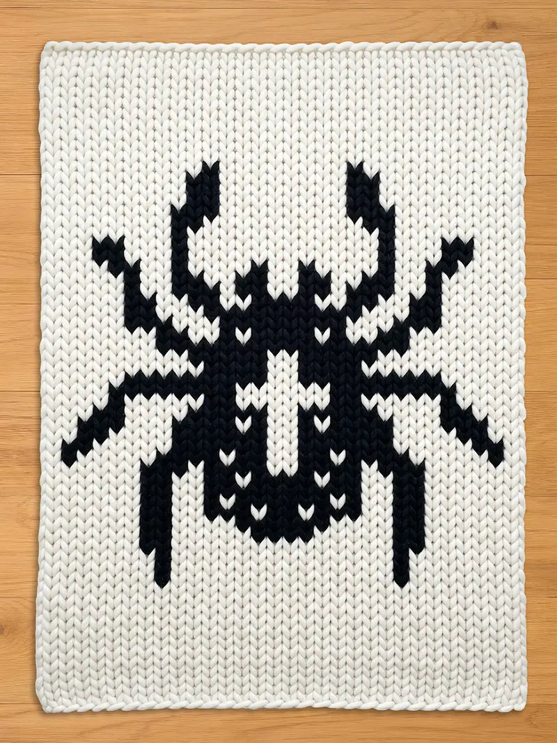 A square, off-white knitted blanket featuring a large, black pixelated insect design. The insect has a segmented body, multiple legs, and antennae. The blanket is made of thick yarn.