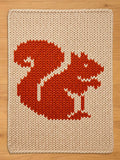 A knitted beige and orange chunky blanket featuring a stylized squirrel design. The squirrel is rendered in orange with white accents, set against a beige background. The blanket has a textured, woven appearance.