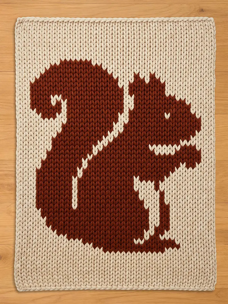 A knitted chunky blanket featuring a brown squirrel design on a cream background. The squirrel is depicted in a simple, pixelated style, with a fluffy tail and a smiling face. The blanket is made of thick yarn.