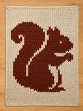 A knitted chunky blanket featuring a brown squirrel design on a cream background. The squirrel is depicted in a simple, pixelated style, with a fluffy tail and a smiling face. The blanket is made of thick yarn.