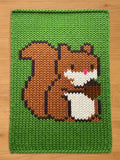 A knitted green chunky blanket featuring a pixelated squirrel design. The squirrel is brown, white, and black with a pink nose. The blanket has a thick, textured appearance, suitable for a child's room or nursery.