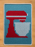 A knitted chunky blanket featuring a red stand mixer with a white bowl. The mixer has white stripes. The background is light blue, and the mat has a white border. The blanket is placed on a wooden surface.