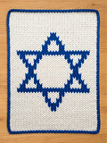A chunky knitted white and blue Star of David design. The central star is blue, with a white background. The border is blue. The item is likely a decorative piece or a small blanket.