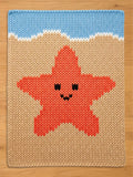 Knitted chunky blanket featuring a smiling orange starfish on a beige background, with a blue sky and white waves at the top. The design is reminiscent of a beach scene, perfect for a child's room or coastal decor.