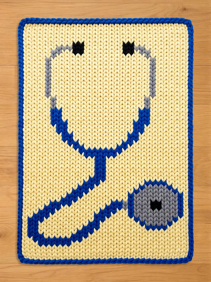 A chunky knit blanket featuring a large blue and grey stethoscope design on a cream background, with a matching blue knitted border