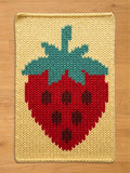 A knitted chunky blanket featuring a large strawberry design. The strawberry is red with dark brown seeds and a green and teal stem. The background is a light yellow color.