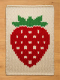 A knitted chunky blanket featuring a large red strawberry design with white seeds and green leaves. The strawberry is centered on a cream-colored background. The blanket has a textured, woven appearance.