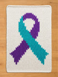 A rectangular white chunky knit blanket laid flat on a light wood floor. Knitted into the center of the blanket is a large awareness ribbon, with the left loop and right tail in teal, and the right loop and left tail in purple