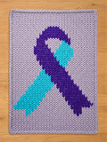 A knitted rectangular blanket featuring a ribbon design. The ribbon is composed of purple and teal sections, set against a light purple background. The blanket is made of thick yarn, with a visible knit texture.