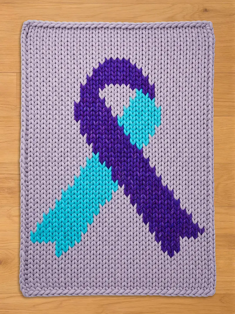 A knitted rectangular blanket featuring a ribbon design. The ribbon is composed of purple and teal sections, set against a light purple background. The blanket is made of thick yarn, with a visible knit texture.
