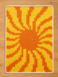 A rectangular, knitted chunky blanket with a sunburst design in shades of yellow and orange. The blanket has a white border and is placed on a wooden surface. The knitted texture is clearly visible, showcasing the craftsmanship of the piece.