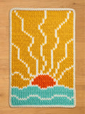 Rectangular chunky knit blanket laid flat, showcasing a vibrant sun and ocean landscape. The knit pattern features a bright orange semi-circle sun, a mustard yellow sky with white jagged sun rays, teal blue water with white wave accents, and a thick white braided border