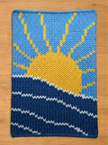 A knitted rectangular chunky blanket featuring a sun and sea design. The upper portion depicts a bright yellow sun with rays against a light blue sky. The lower section shows dark blue waves with white accents, creating a nautical theme.